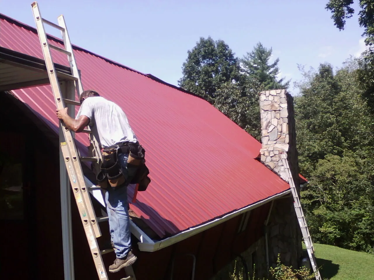 Roofer on a ladder beside a red metal roof and stone chimney — Soffit & Fascia Repair by Roofing Brothers in Romeoville